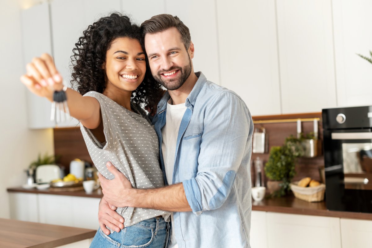 Young interracial married couple homeowners smiling, showing keys from a new apartment, hugging and looking at the camera, standing in the kitchen and celebrating moving in a new home, family concept