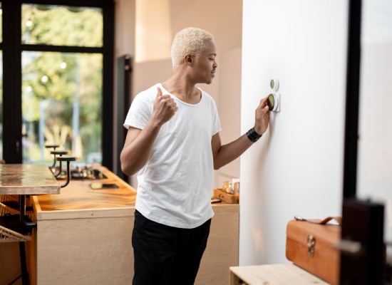 Latin man choosing temperature on thermostat. Young focused guy pushing button on smart home system. Concept of modern domestic lifestyle. Interior of kitchen in modern apartment.