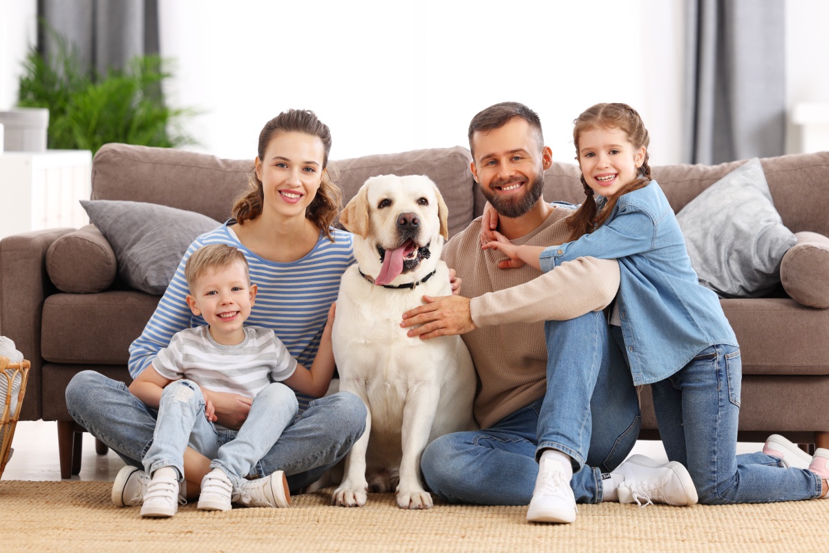 Smiling mother and father with little kids and adorable purebred Labrador retriever dog sitting on floor and looking at camera while spending time together at home